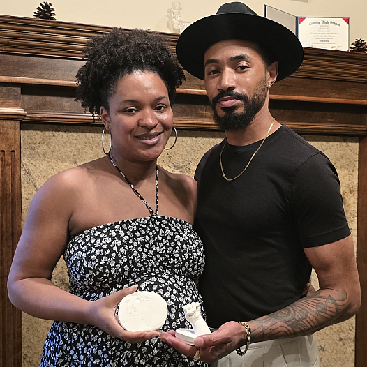 A Black couple stands in front of a wooden mantle holding plaster molds of their late daughters hand and foot.
