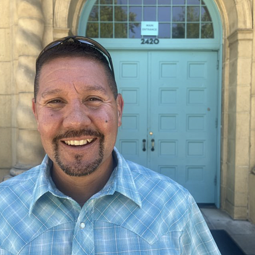 J Vasquez stands in a blue shirt smiling in front of blue double doors.