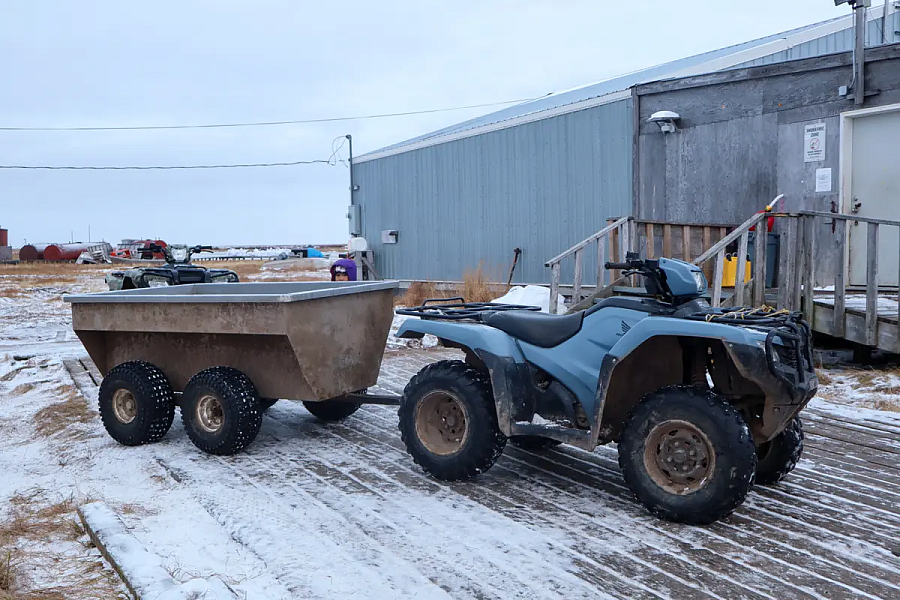 A four-wheeler ATV with a trailer, known locally as the “bathroom bus,” is parked outside a rural Alaskan school building on a snowy wooden boardwalk.