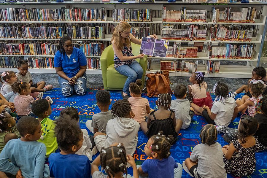 A lady sitting on a couch reads a book to kids sitting on the ground