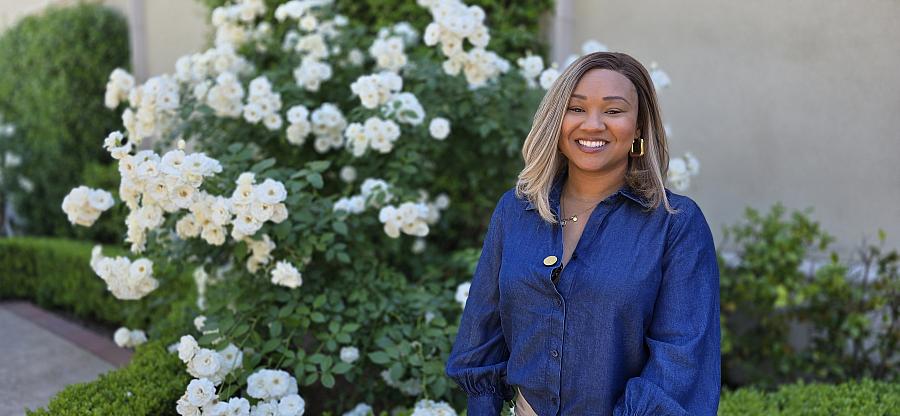 A young Black woman sits in a white rose garden smiling in a blue denim shirt.
