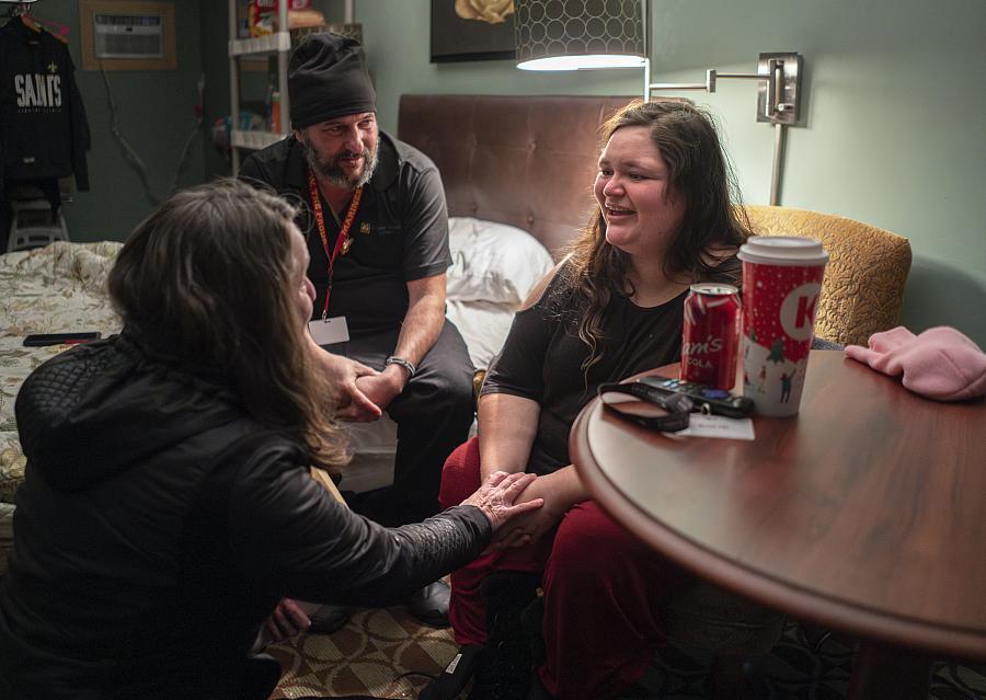 A woman smiles while speaking with two others in a cozy room. One person holds her hands supportively. A soda, coffee cup, and keys rest on the nearby table.