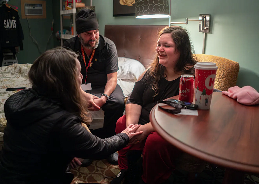 Three people sitting inside a room, one besides a table, one on a bed talking to each other.