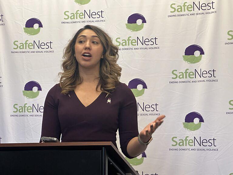 A woman speaking at a press conference with SafeNest poster behind her.