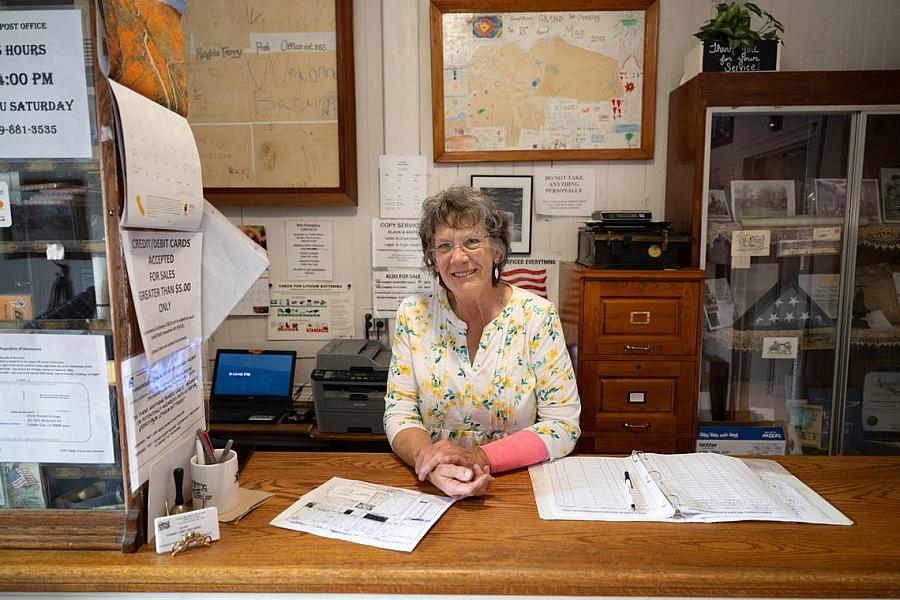 A smiling woman stands behind the counter of a small post office or local office, surrounded by papers, a binder, a computer, and various notices and decorations on the wall