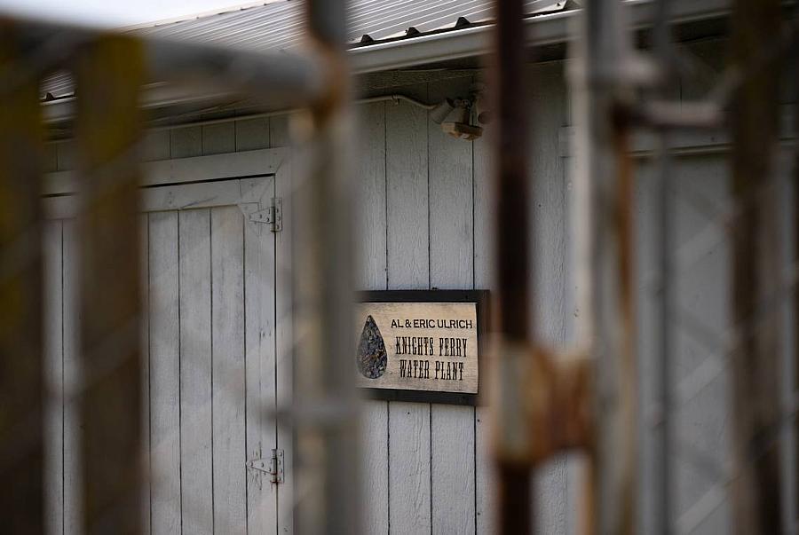 A close-up view of a wooden building with a sign reading ‘Al & Eric Ulrich Knights Ferry Water Plant,’ partially obscured by a chain-link fence in the foreground