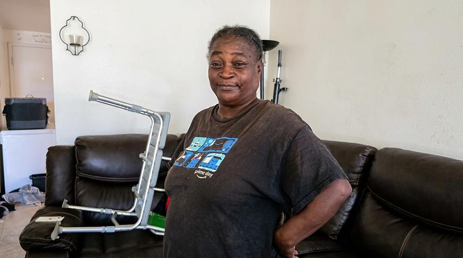 An older Black woman stands in her Shafter, California living room by the shower bench her granddaughter Sarabi uses to bathe. 