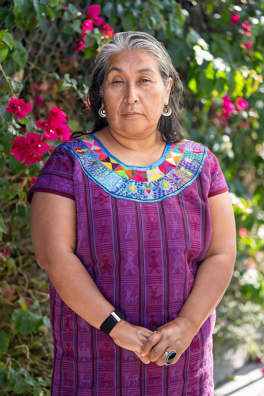 Odilia Romero with grey hair stands in front of colorful flowers in a purple dress.