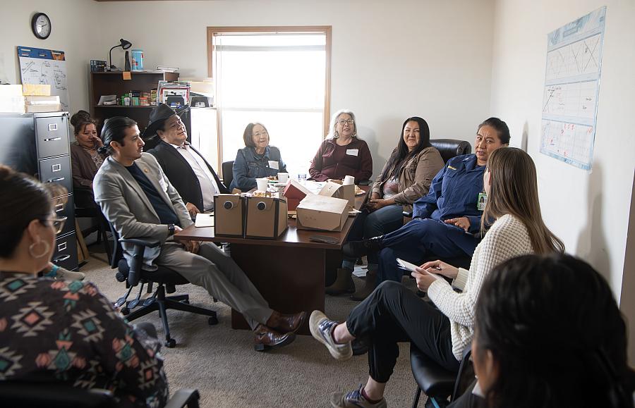 A group of people seated in a circle in a small office space, engaged in a discussion. Some have name tags, and food boxes are placed on the table in the center. A woman in a blue uniform speaks while others listen attentively.