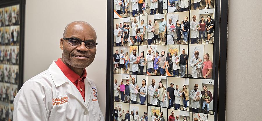 A bald Black doctor stands in front of all the mothers and babies he has helped bring into the world,