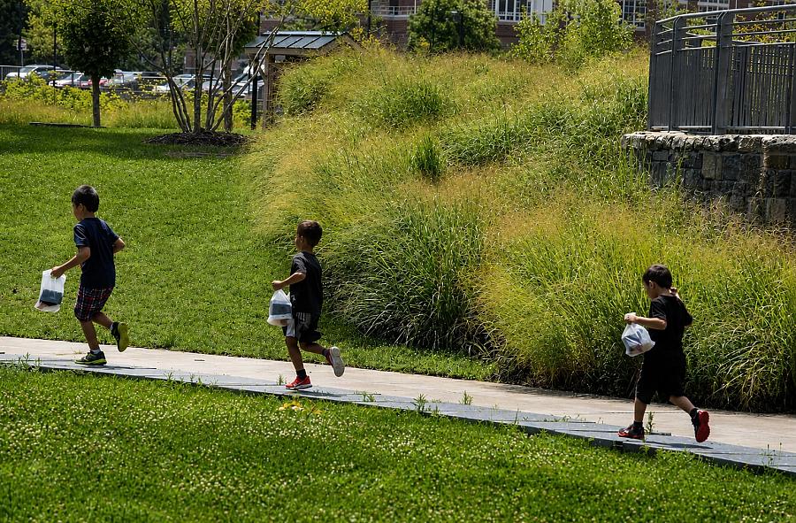 Three children run through a park with bags in their hands.