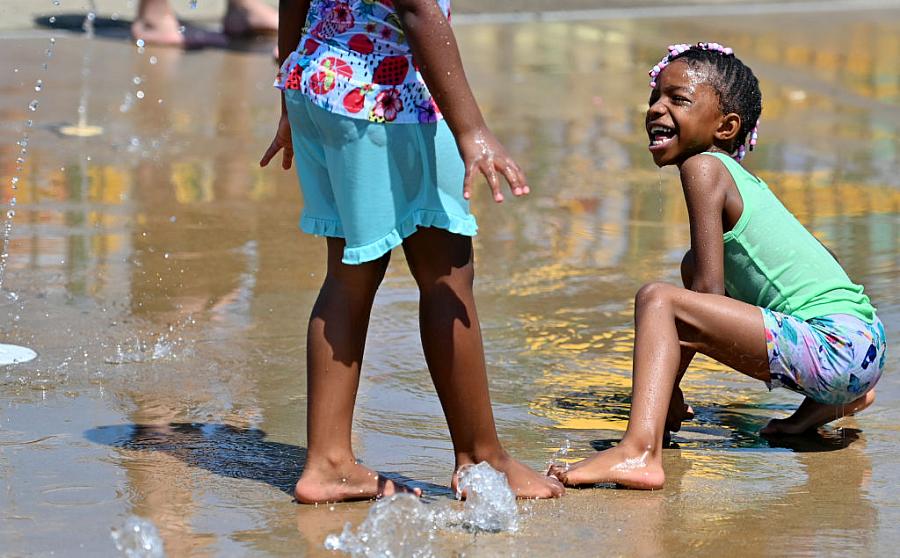 A child plays with a friend in a sprinkler.