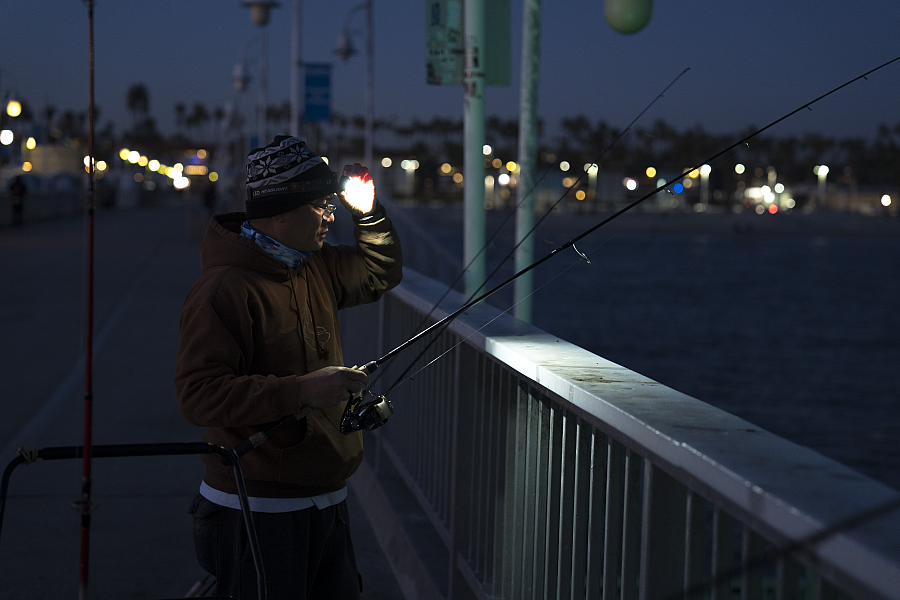 A person wearing a brown hoodie and knit hat is night fishing on a pier, illuminated by a headlamp. They are holding a fishing rod and standing by a railing, with the ocean and city lights visible in the background.
