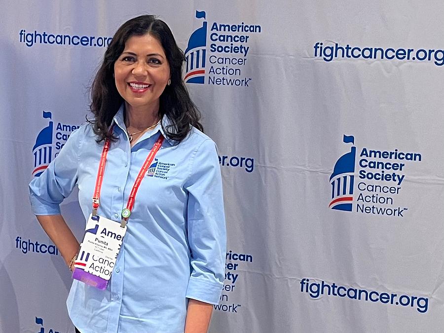 A young smiling Indian woman stands in scrubs in front of a step-and-repeat background at a cancer event