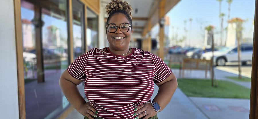 A young smiling Black woman wears a striped red and white t-shirt and stands outside on the sidewalk.