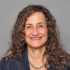 woman with brown curly hair smiling wearing a pearl necklace and dark blazer