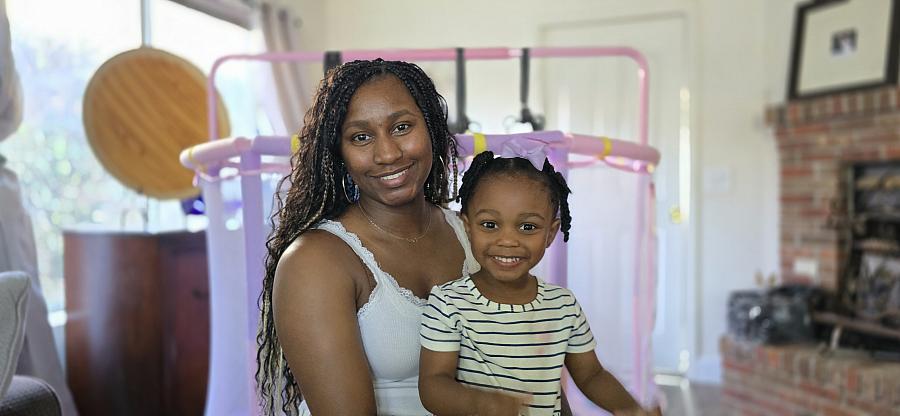 A young Black woman sits smiling with her daughter, also smiling, on her lap.