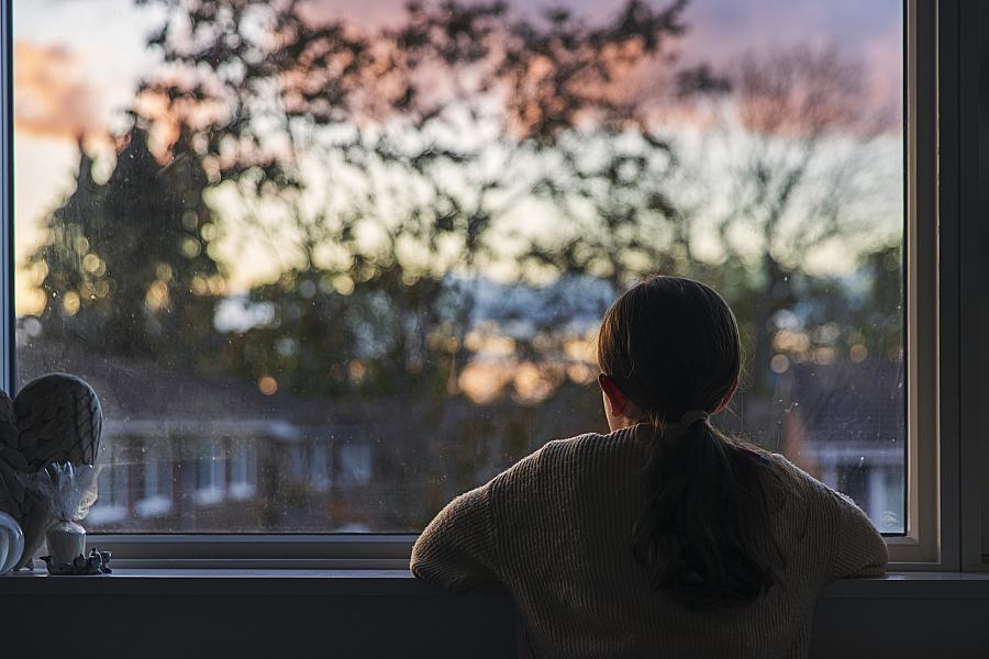 Person staring outside window at a scenery of tress and houses with twilight in the background