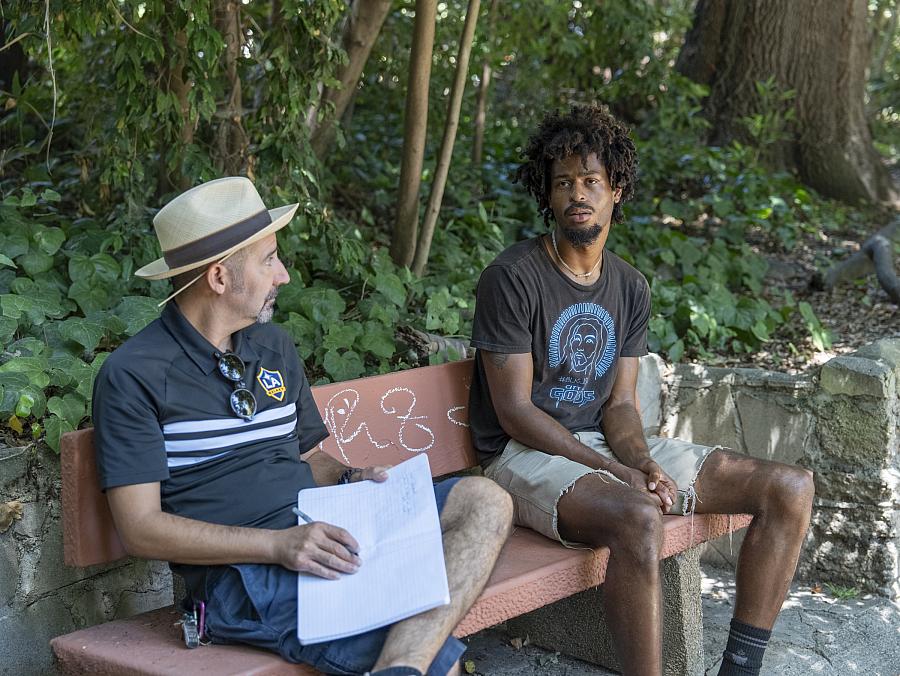 A young man sits on a park bench speaking with an older man who holds a notebook and pen. They appear to be engaged in a serious conversation surrounded by greenery.
