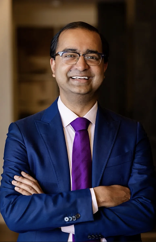 An Indian man in a dark blue suit and glasses smiles in his office