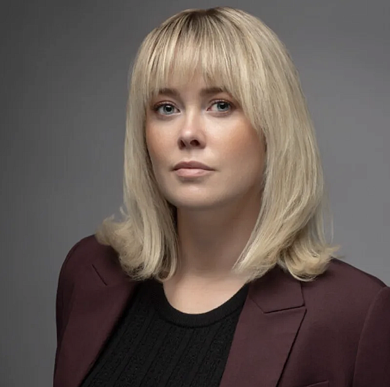 A white blond woman with bangs and a burgundy blazer looks at the camera 