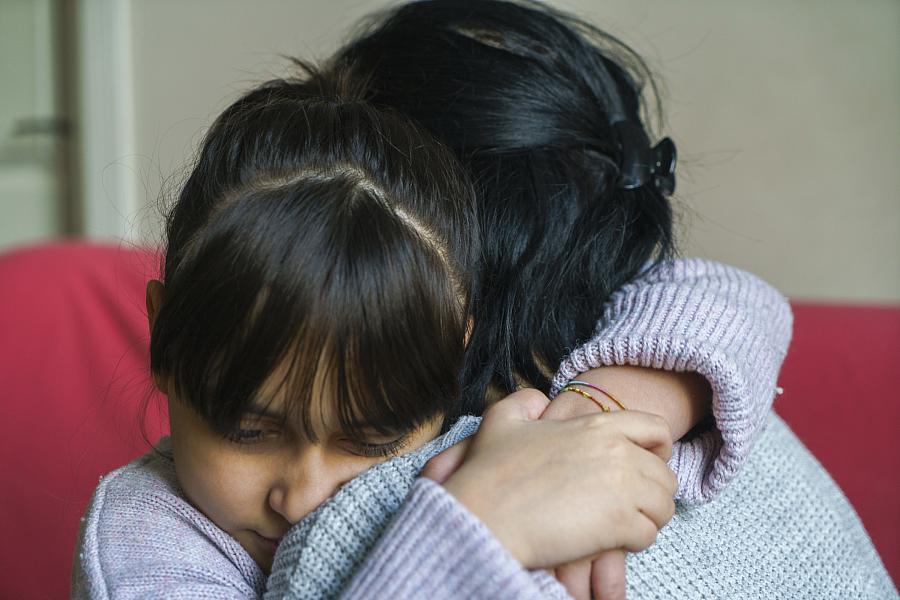 A young girl with bangs hugs an adult tightly, both wearing sweaters, in an emotional moment of comfort and support.