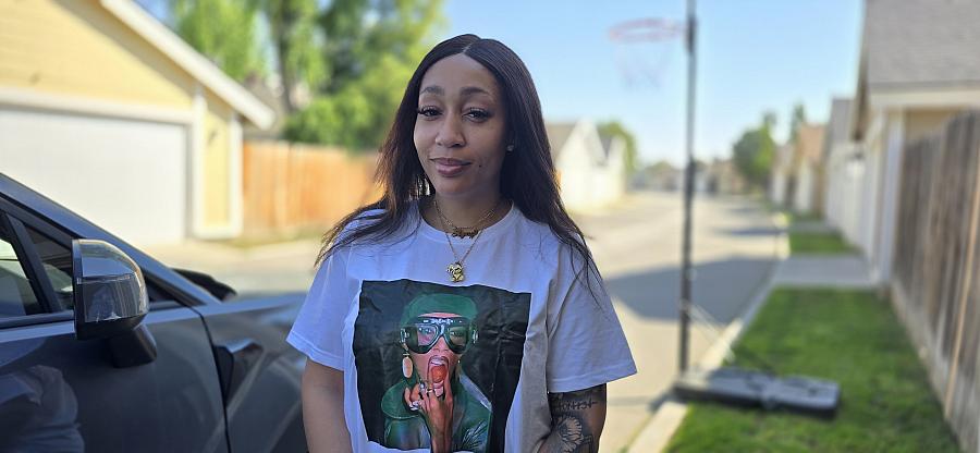 A young Black women wears a white t shirt and stands outside on the street beside a car and a basketball hoop.