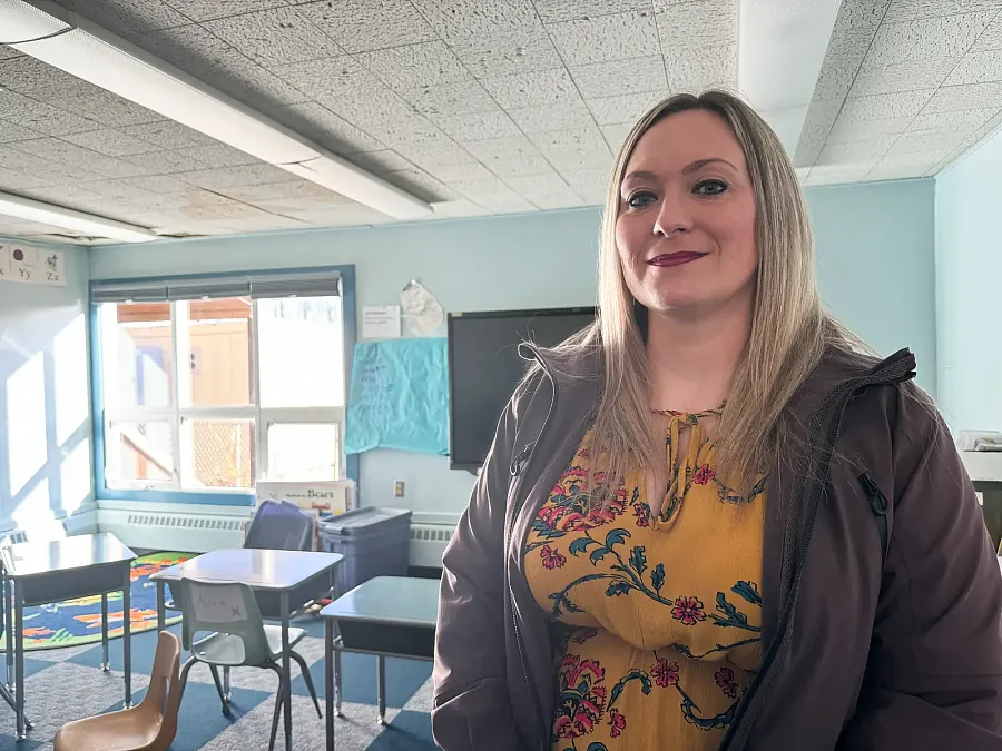 A teacher stands inside a brightly lit but aging rural Alaskan classroom with missing ceiling tiles and worn furniture.