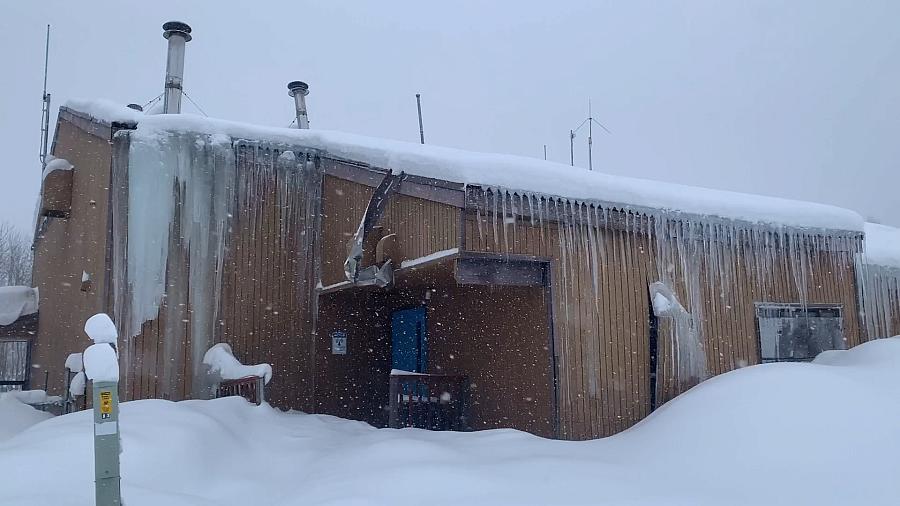 A snow-covered building with large icicles hanging from the roof, showing signs of structural damage and surrounded by deep snow.