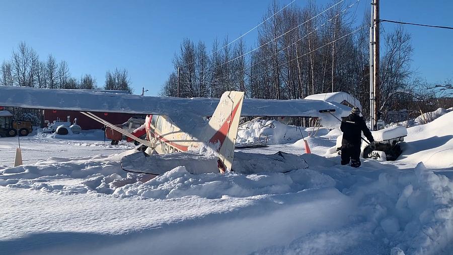 A small airplane buried in deep snow appears damaged, with a person nearby inspecting the scene on a sunny winter day in a rural area.
