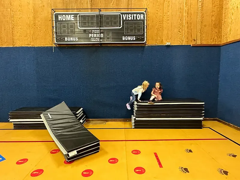 Two young children play on stacked gym mats under a scoreboard in a school gym with colorful floor markings.