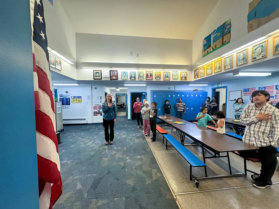 Students and staff stand with hands on hearts, facing the American flag for the Pledge of Allegiance in a school hallway decorated with portraits and lockers.