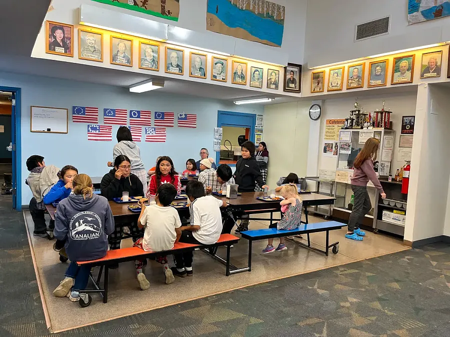 Students eat and socialize at lunch tables in a school cafeteria decorated with portraits and American flags.