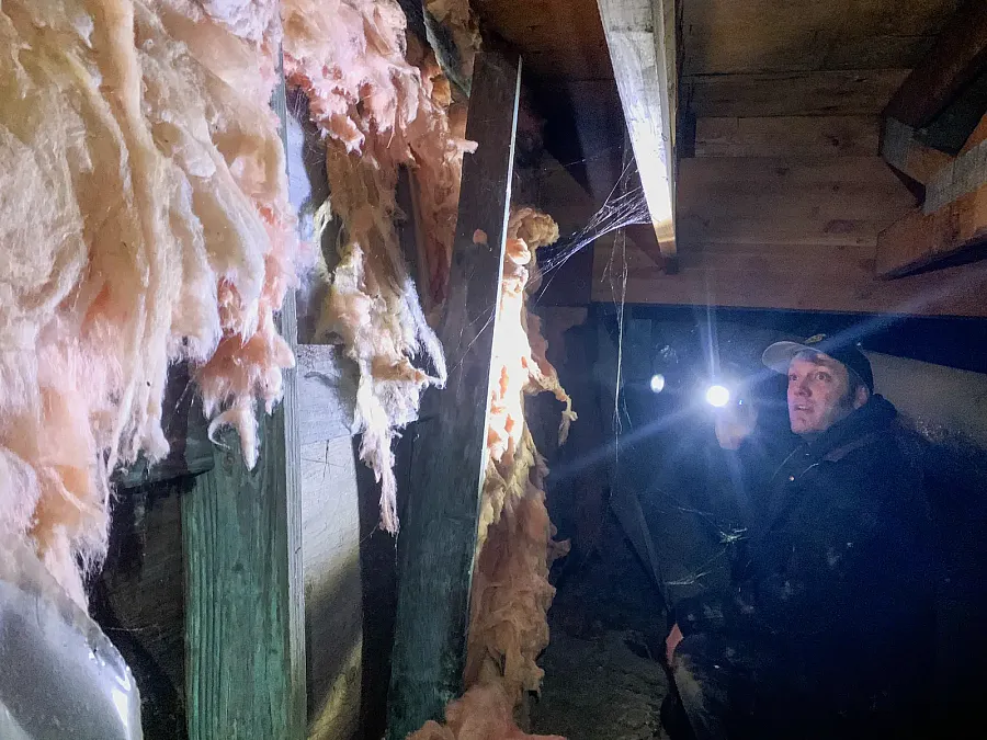 A man crouches with a flashlight in a dark crawlspace beneath a school, examining sagging insulation and cobweb-covered wooden beams, highlighting the building’s deteriorating condition.
