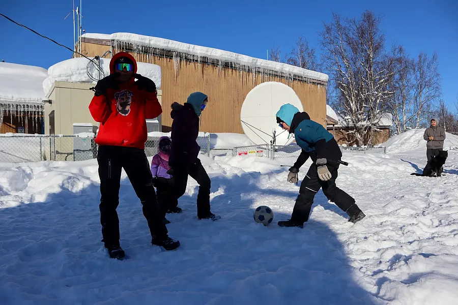 Children bundled in winter clothing play soccer in deep snow outside a snow-covered school building in Sleetmute, Alaska, with icicles hanging from the roof and a dog in the background.