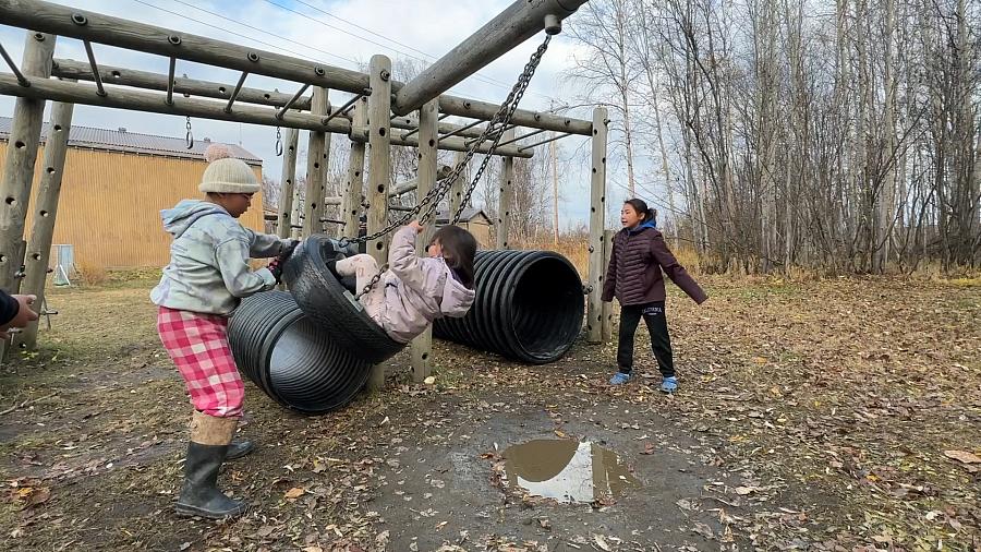 Children play on a tire swing and climbing structure in a muddy, leaf-covered playground near a rural Alaskan school, surrounded by bare trees and wooden buildings.
