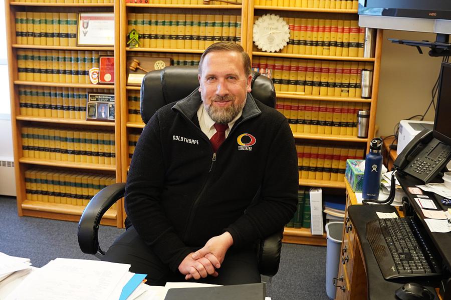 A man sitting in an office lined with legal books, wearing a black jacket labeled “Goldthorpe” and smiling at the camera, possibly a public official or legal professional.