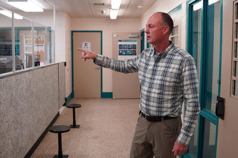 A man in a checkered shirt pointing while giving a tour inside a detention facility or institutional hallway