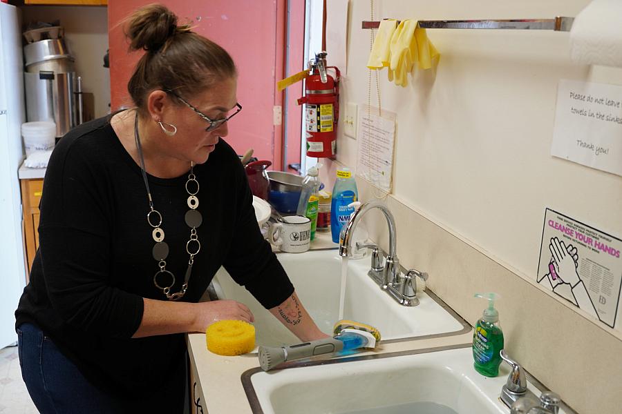 A woman washing dishes at a sink in a communal or institutional kitchen, with various cleaning supplies visible.