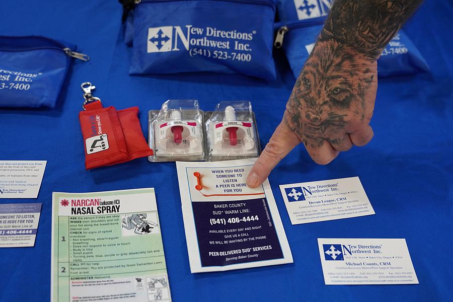 A hand pointing to a card on a table covered with materials related to overdose prevention, including Narcan nasal spray and peer support contact information.