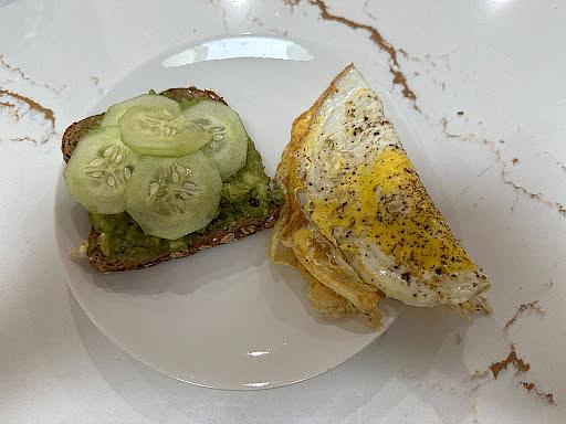A white plate shows a piece of toast with cucumber alongside one fried egg.