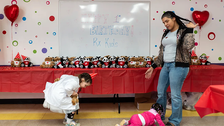 A young girl in a party hat looks at a crawling baby in front of a display of teddy bears and a birthday sign for “Rx Kids.