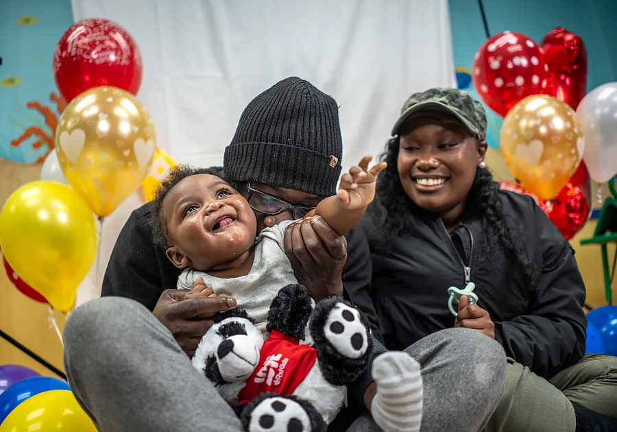 A smiling toddler is embraced by an adult while another adult looks on, surrounded by balloons and stuffed pandas.