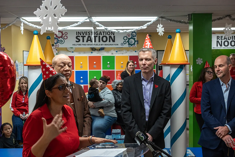 A woman speaks at a podium during a celebration, with party hats, colorful decor, and people gathered behind her.