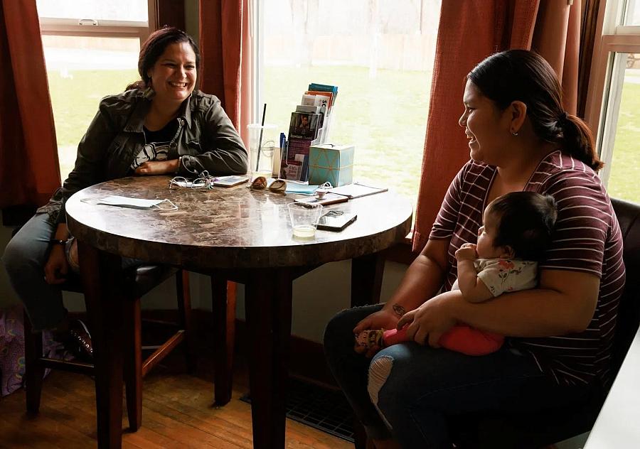 Two women speaking to each other, sitting across table, one of them is holding child