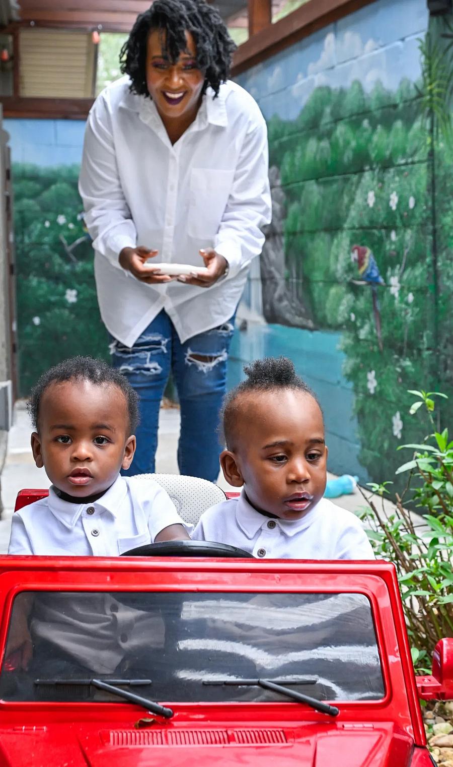 person standing behind 2 children in play car