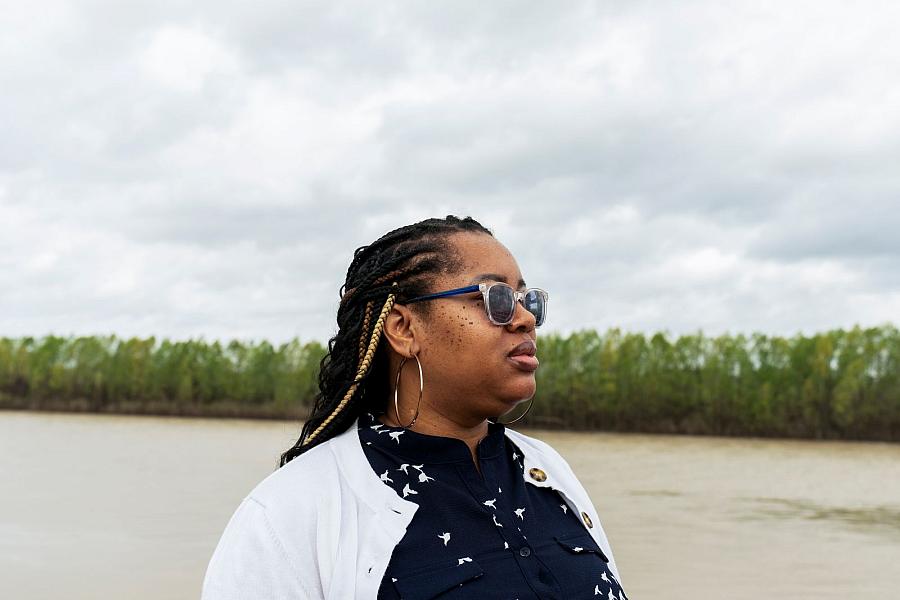 Woman with braids and glasses stands near a river under a cloudy sky.
