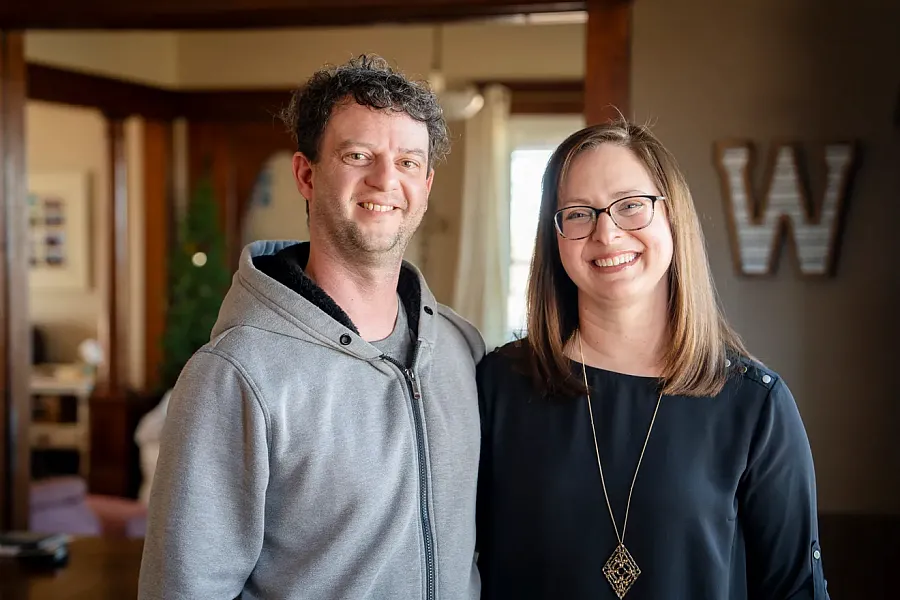 Smiling man in a gray hoodie and woman in glasses and a black top stand together in a warmly lit home interior.
