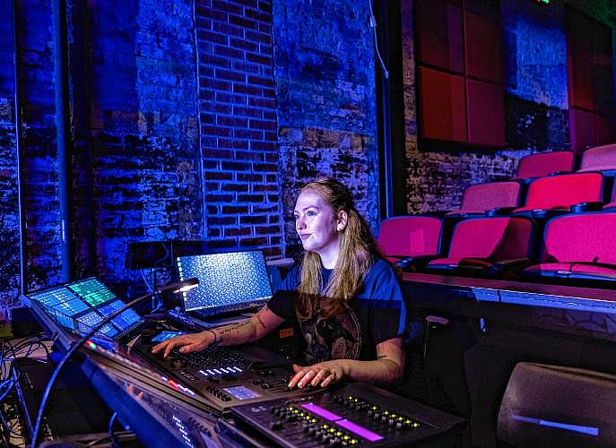 A woman working at a sound and lighting control panel in a dark theater with blue lighting.