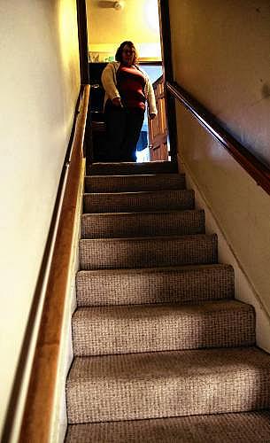 A woman stands at the top of a narrow, carpeted staircase in a dim hallway.